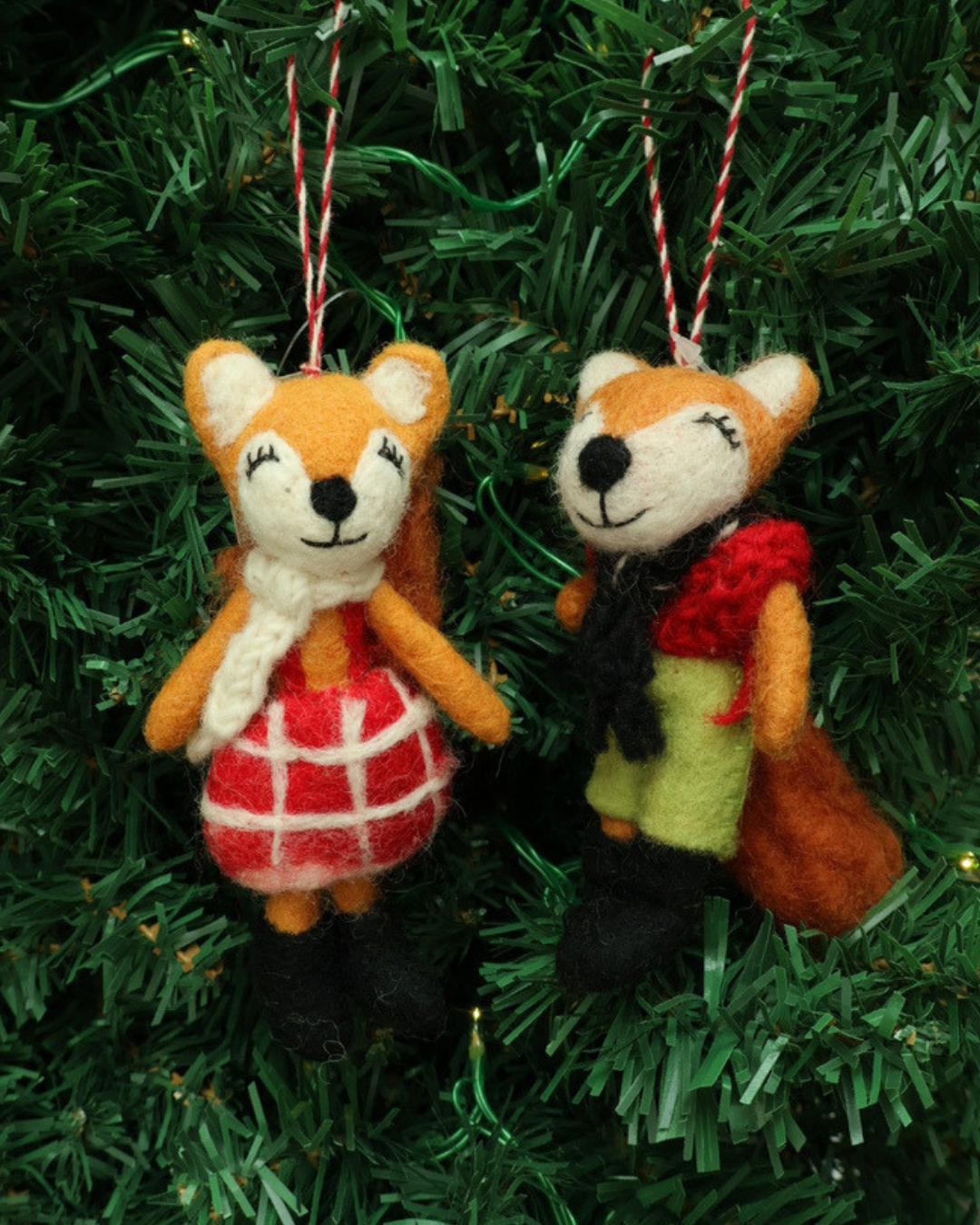 Dekorierter Weihnachts-Fuchs-Filzanhänger "Naomi und Nils" von Grace & Gloria hängend an einem Tannenbaum. Es sind 2 süße Füchse mit einer grünen Hose und einem roten Kleid.