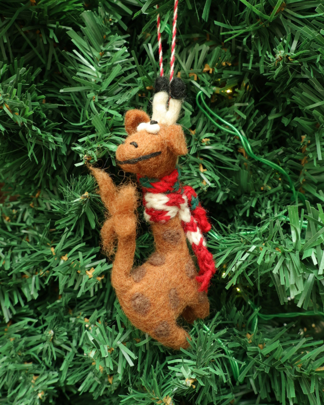 Dekorierter Giraffen Weihnachts-Filzanhänger "Willma" von Grace & Gloria hängt an einem Tannenbaum. Die Giraffe ist braun mit dunklen Flecken, trägt einen rot/weißen Schal und schaut nah hinten.