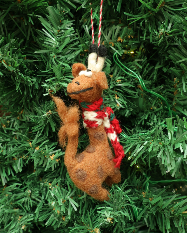 Dekorierter Giraffen Weihnachts-Filzanhänger "Willma" von Grace & Gloria hängt an einem Tannenbaum. Die Giraffe ist braun mit dunklen Flecken, trägt einen rot/weißen Schal und schaut nah hinten.