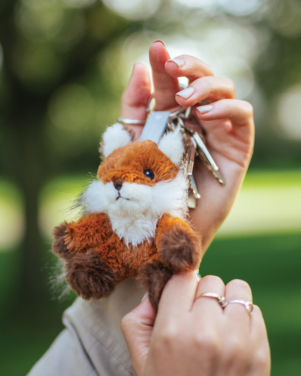 Fuchs Schlüsselanhänger Autumn von Wrendale Designs, an einem Schlüsselring in einer Hand gehalten, mit rostfarbenem Kuschelfell, weißer Schnauze und dunklen Pfoten vor unscharfem grünem Hintergrund im Freien.