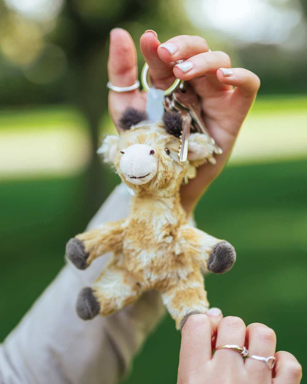 Giraffen Schlüsselanhänger Camilla von Wrendale Designs, an einem Schlüsselring in einer Hand gehalten, mit braunem Kuschelfell und kleinen Hörnchen vor unscharfem grünen Hintergrund im Freien.