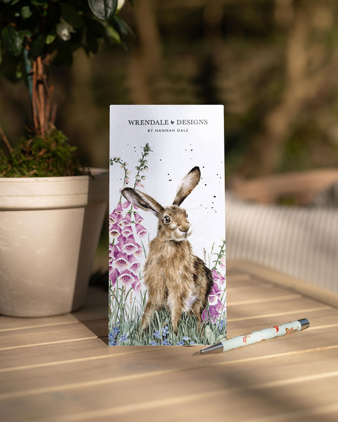 Hasen Notizblock "The WIld Garden" von Wrendale Designs  auf einem Holztisch im Garten. Das Motiv ist ein Hase auf einer Blumenwiese welcher in die Kamera schaut. 