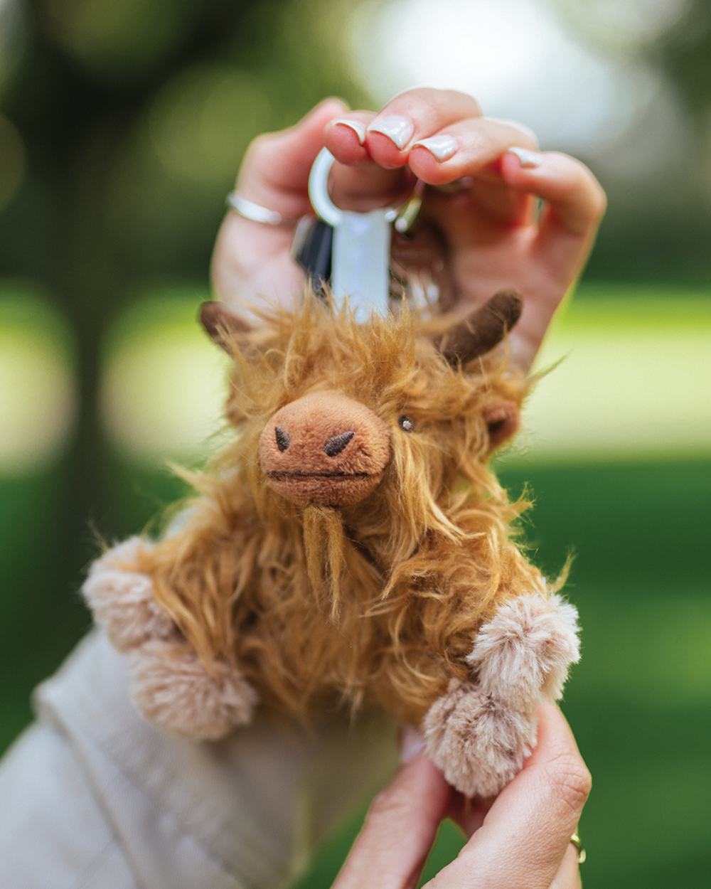 Hochland Kuh Schlüsselanhänger Gordon von Wrendale Designs, an einem Schlüsselring in einer Hand gehalten, mit zotteligem braunem Fell und kleinen Hörnern vor unscharfem grünen Hintergrund im Freien.