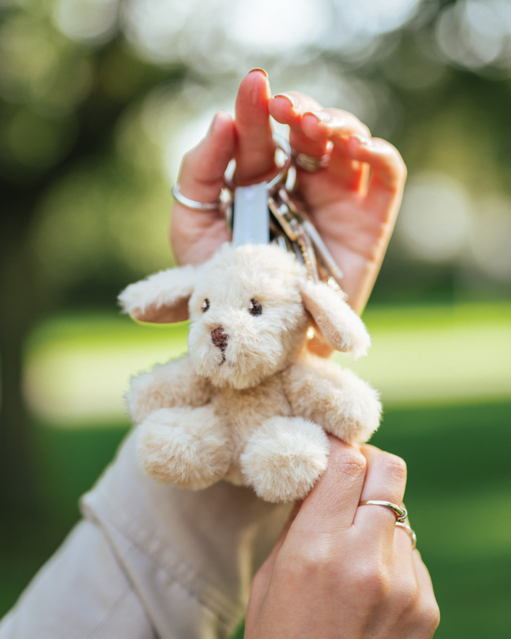 Hunde Schlüsselanhänger Ralph von Wrendale Designs, an einem Schlüsselring in einer Hand gehalten, mit hellem Kuschelfell und Schlappohren vor unscharfem grünem Hintergrund im Freien.