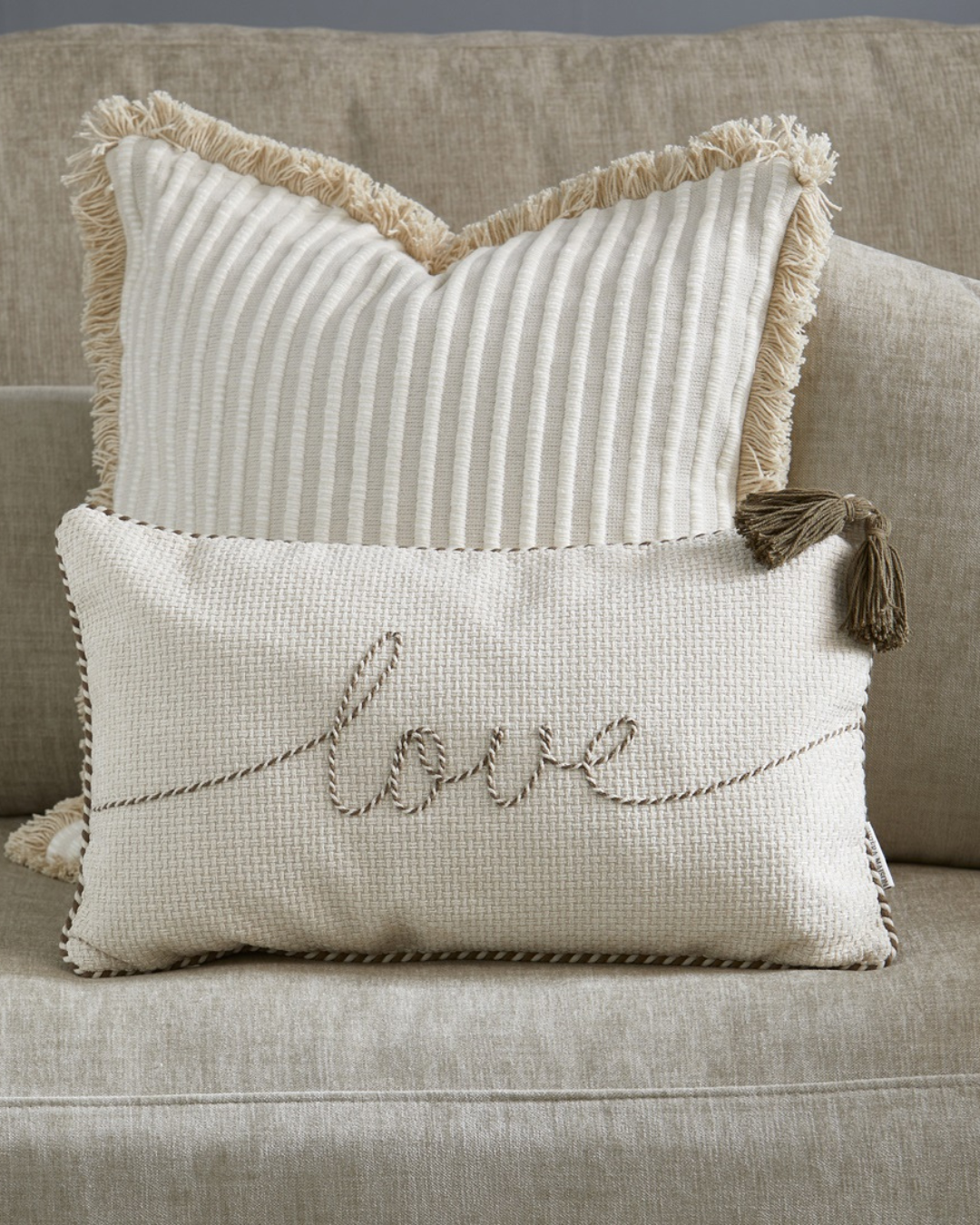 Dekorativer Kissenbezug „With Love Pillow Cover 50x30“ von Rivièra Maison mit gesticktem Schriftzug „love“, liegend vor einem größeren hellen Kissen auf einem Sofa mit Stoffbezug.