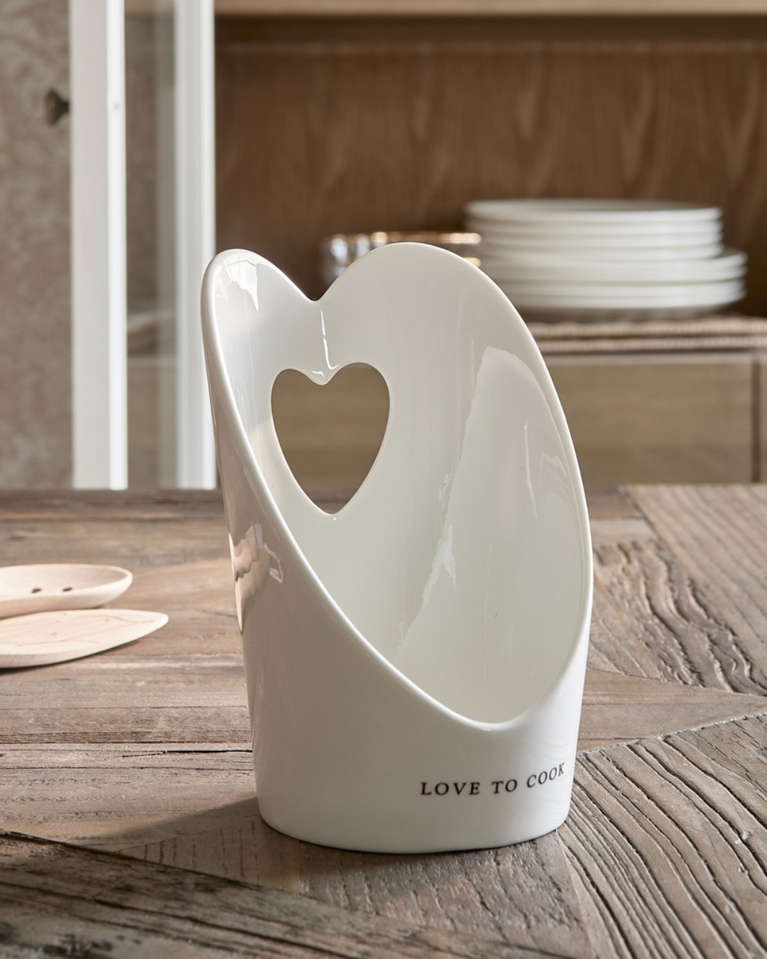 Weißer Kochlöffelhalter „With Love Spoon Holder“ von Rivièra Maison auf rustikalem Holztisch in einer Küche, Herzform und Schriftzug gut erkennbar.