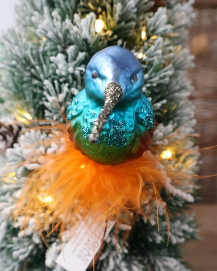 Exotisches Kolibri Ornament zum Klemmen von Inge Glas vor dekoriertem Tannengrün. Der aus Glas gefertigte Kolibri sitzt auf echten orangefarbenen Federn. Der Vogel selbst hat orange schimmernde Flügel mit orangefarbenen Glitzer Highlights und blaue Brust- und Kopffedern mit blauen Glitzer Highlights.