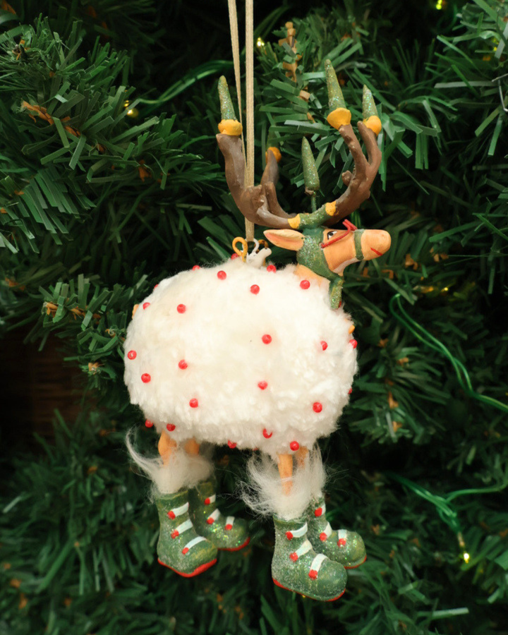 "Dash Away Blitzen Rentier" Krinkle von Patience Brewster hängt an einem Tannenbaum. Zu sehen ist ein Rentier mit rot bepunkteten Schafsfell vor weißen Hintergrund. Daas Rentier hat viele grüne und rote Details.