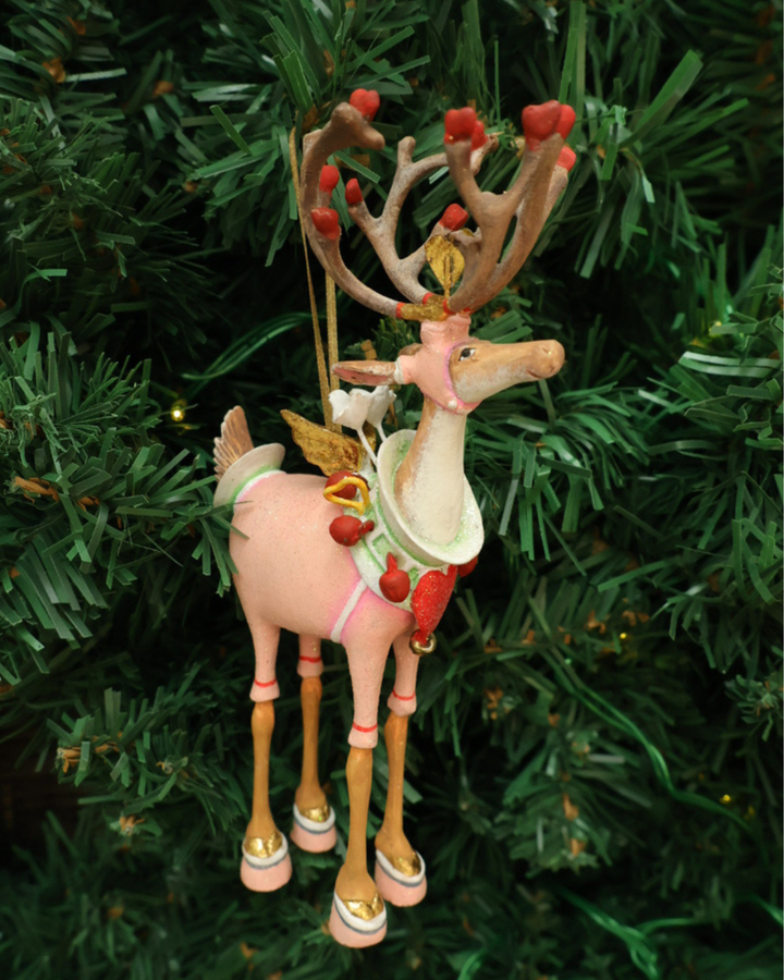 "Dash Away Cupid Rentier" Krinkle von Patience Brewster hängt an einem Tannenbaum. Zu sehen ist ein farbenfrohes Rentier Krinkle mit vielen pinken und roten Detais.