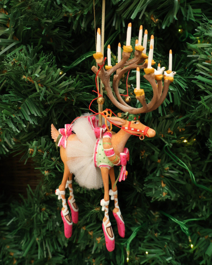 "Dash Away Dancer Rentier" Krinkle von Patience Brewster hängt an einem Tannenbaum. Zu sehen ist 1 farbenfrohes Rentier Krinkle welches ein weißes Rock trägt mit vielen pinken und weißen Details.