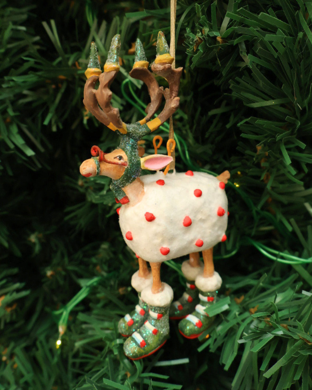 Mini "Dash Away Blitzen Rentier" Krinkle von Patience Brewster vor hängt an einem Tannenbaum. Zu sehen ist ein Rentier mit rot bepunkteten Schafsfell vor weißen Hintergrund. Daas Rentier hat viele grüne und rote Details.