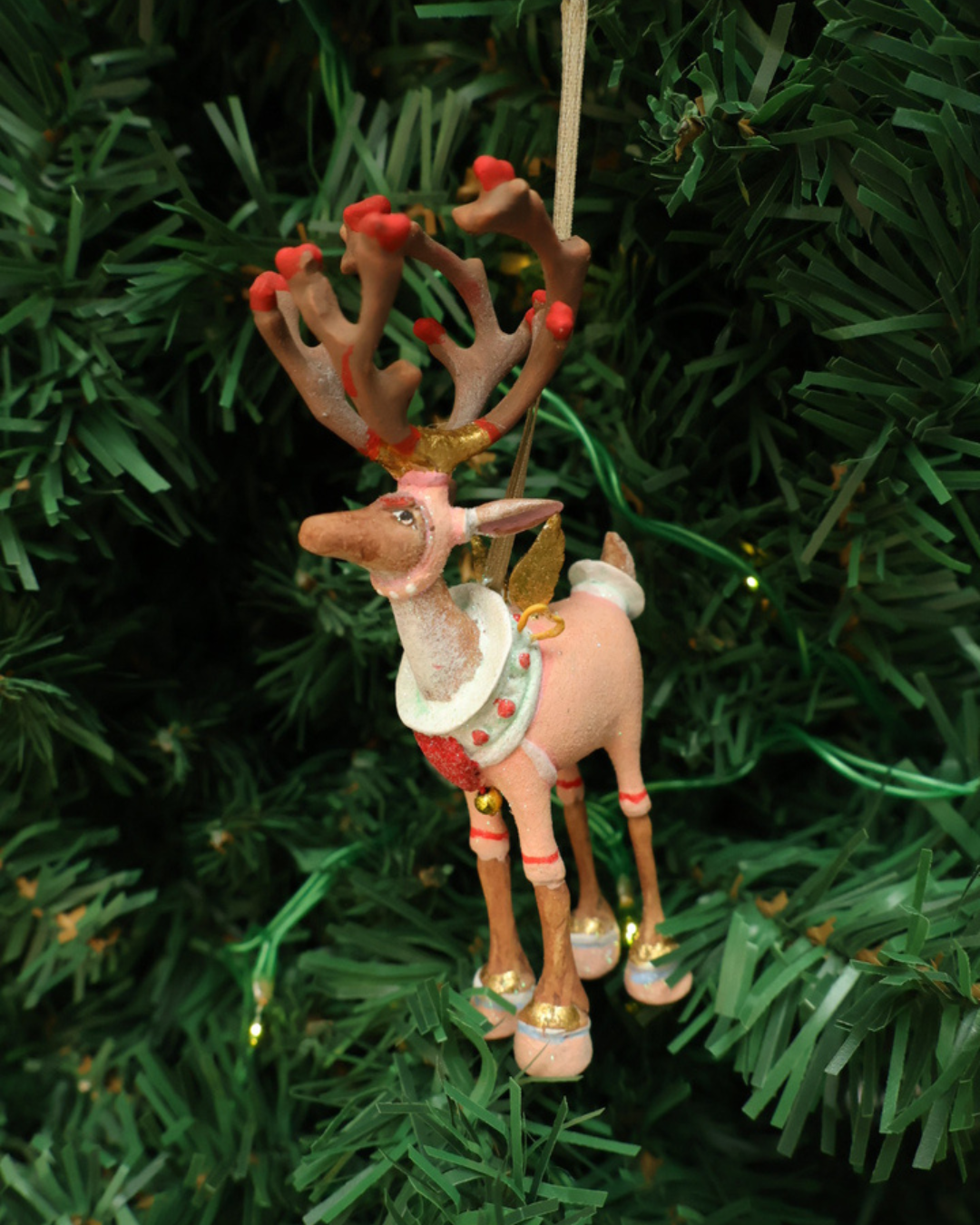 "Dash Away Cupid Rentier" Krinkle von Patience Brewster hängt an einem Tannenbaum. Zu sehen ist ein farbenfrohes Rentier Krinkle mit vielen pinken und roten Detais.