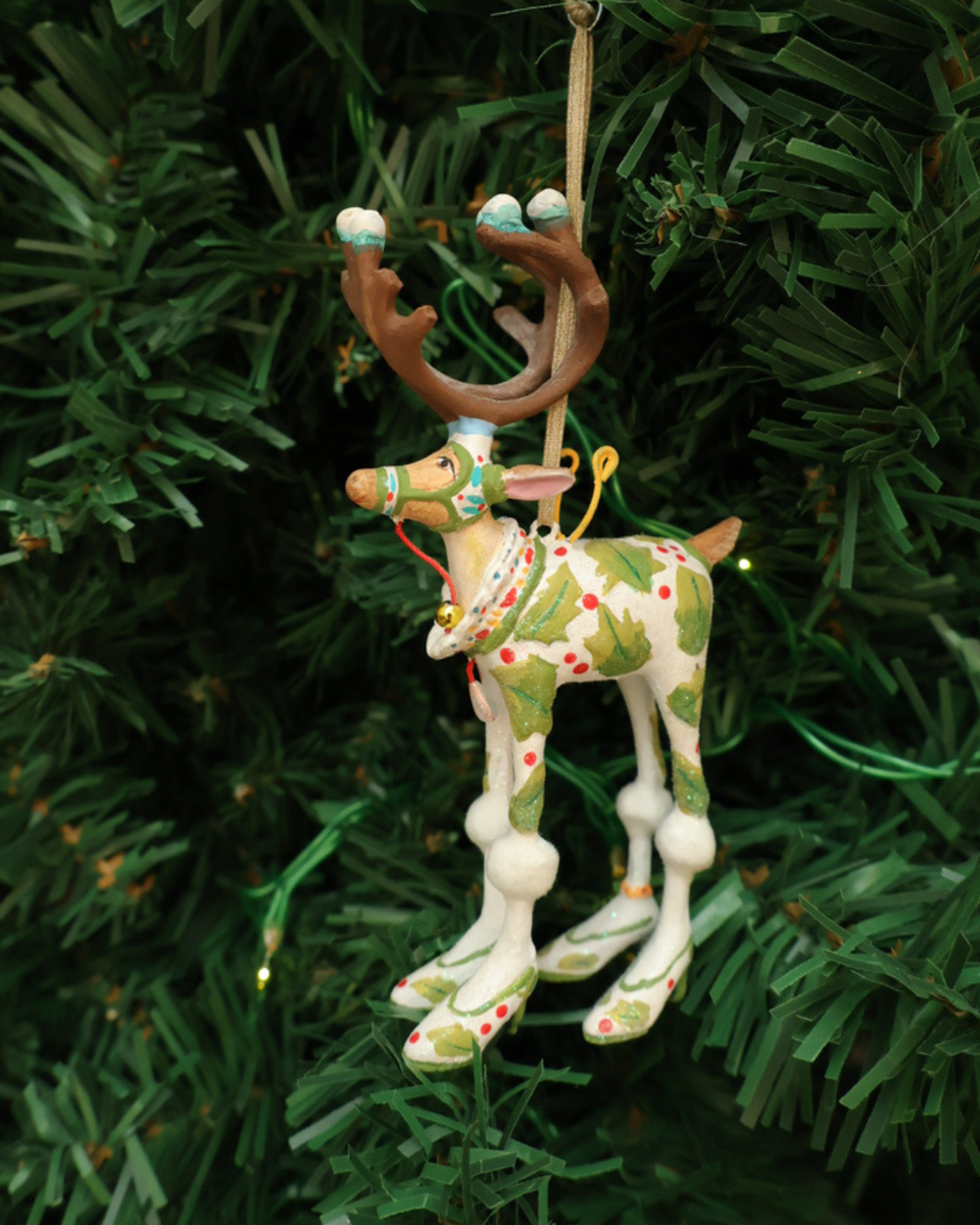"Dash Away Vixen Rentier" Krinkle von Patience Brewster hängt an einem Tannenbaum. Zu sehen ist ein farbenfrohes Rentier Krinkle mit vielen grünen und weißen Detais.