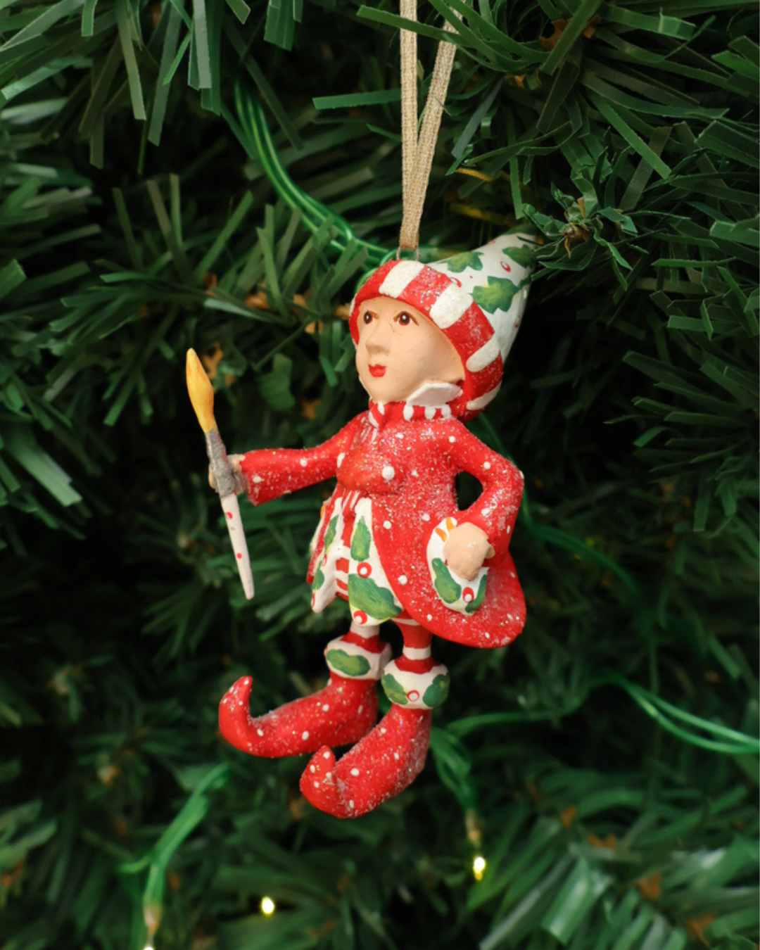 Mini "Vixens Elf" Krinkle hängt an einem Tannenbaum. Zu sehen ist ein größtenteils rot bekleideter Elf mit weißen Pünktchen, pflanzlichen Details und einem kleinen Pinsel in der Hand. 