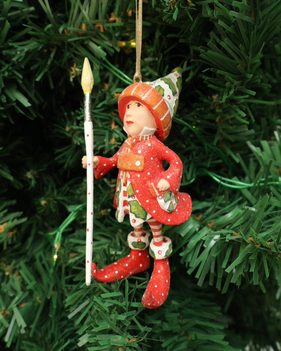 "Vixens Elf" Krinkle hängt an einem Tannenbaum. Zu sehen ist ein größtenteils rot bekleideter Elf mit weißen Pünktchen, pflanzlichen Details und einem großen Pinsel in der Hand. 