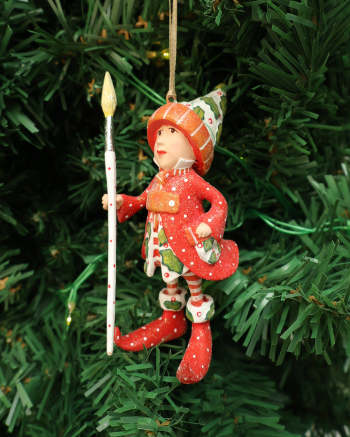"Vixens Elf" Krinkle hängt an einem Tannenbaum. Zu sehen ist ein größtenteils rot bekleideter Elf mit weißen Pünktchen, pflanzlichen Details und einem großen Pinsel in der Hand. 