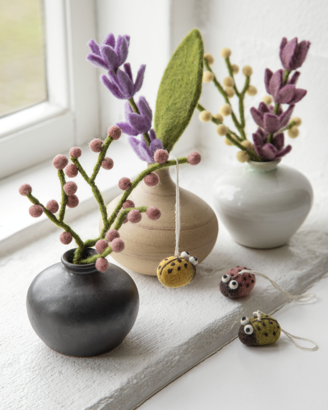 Rougefarbener Mini Beeren-Ast aus Filz von Gry & Sif kombiniert mit Filzblumen in Vasen auf einer Fensterbank, helles Tageslicht, ruhige Frühlingsszene mit Keramikvasen.