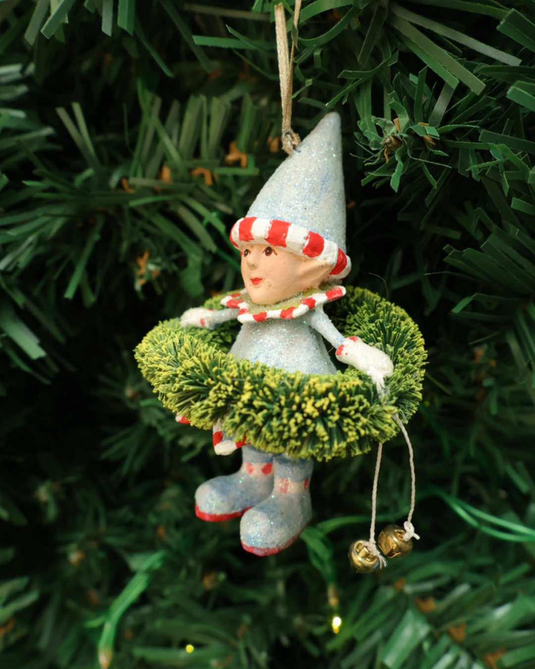 Mini "Dashers Elf" Krinkles von Patience Brewster hängt an einem Tannenbaum. Zu sehen ist ein Elf mit hellblauen Kleid, Hut, Schuhen und grünen Pflanzen Details. Der Elf hält 2 Glocken in der Hand.