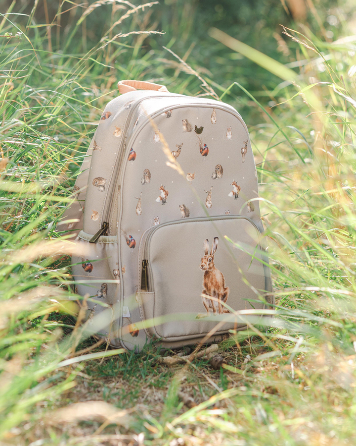 Rucksack „Woodlanders“ von Wrendale Designs steht im hohen Gras, grauer Stoff mit Waldtiermotiven und Hasenillustration auf der Fronttasche.