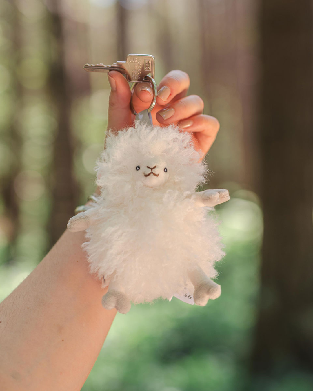 Flauschiger Schaf Schlüsselanhänger Beryl von Wrendale Designs, an einem Schlüsselring in einer Hand gehalten, vor unscharfem Waldhintergrund im Freien.
