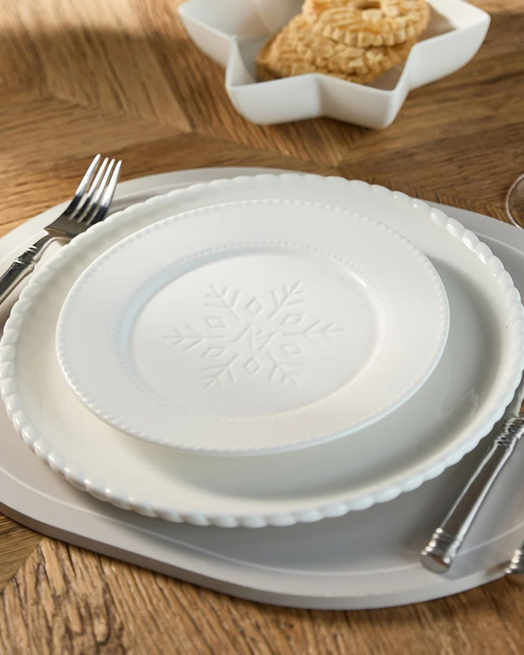 Weihnachtlicher weißer Frühstücksteller "Snowflake" von Riviera Maison mit feiner Schneeflockenverzierung in der Tellermitte. Im Gesamtensemble mit mehreren Tellern und Besteck auf einem Holztisch platziert.