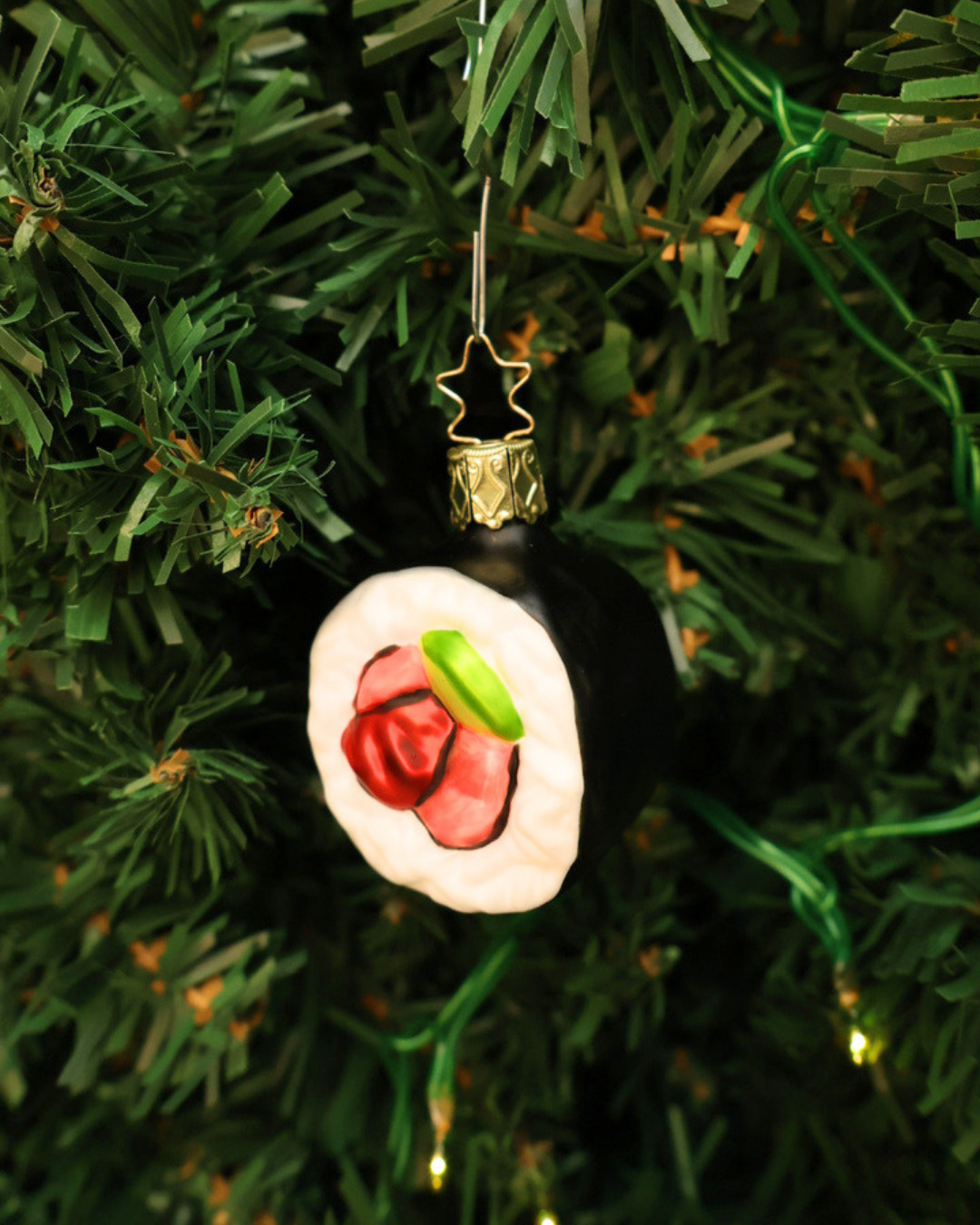 Handgefertigter Sushi Weihnachtsbaumschmuck von Inge Glas mit rosafarbenem Lachs, grüner Avocado, weißem Reis und schwarz-grünem Noriblatt. Hier hängend in einer Tanne angebracht.