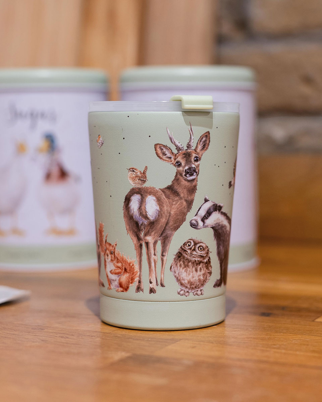 Der Waldtiere Thermobecher To Go "Woodland" von Wrendale Designs ist in hellem grün gestaltet. Detailreiche Waldtiere wie Dachs, Eule, Reh, Eichhörnchen und Zaunkönig illustriert von Hannah Dale zieren den praktischen To Go Becher. Das Foto zeigt den Becher auf Holzboden stehend mit größeren zwei Wrendale Designs Dosen im Hintergrund.
