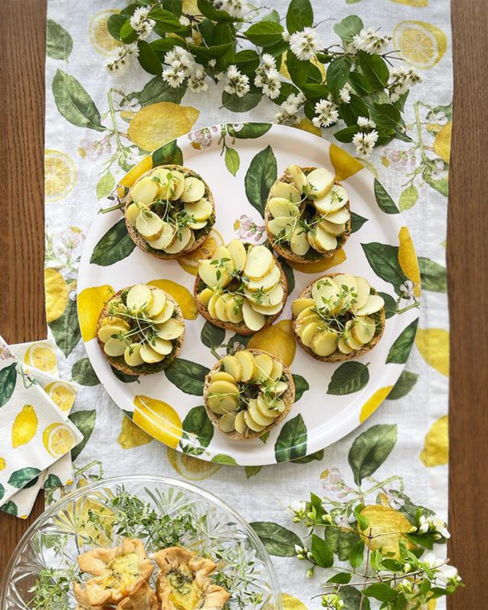 Tischläufer "Zitrone" von Koustrup & co auf hellem Holztisch. Auf dem Tisch liegen Blumen, Teller, Servietten und Speisen welche farblich mit harmonieren.