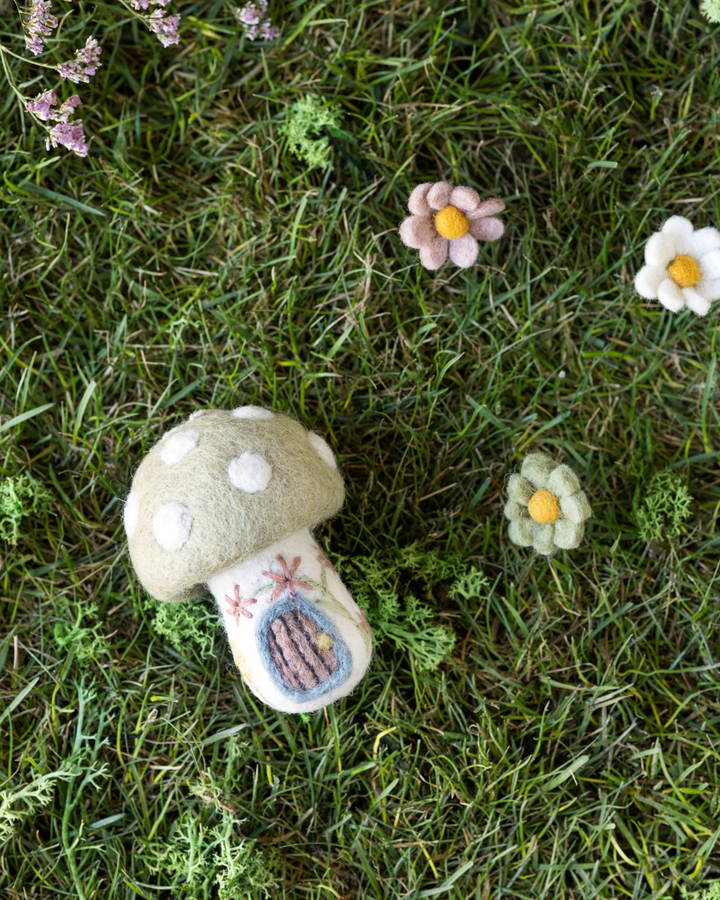 Dekoratives Filz-Pilzhaus von Gry & Sif auf grünem Gras liegend, umgeben von kleinen Filzblumen in Frühlingsfarben.