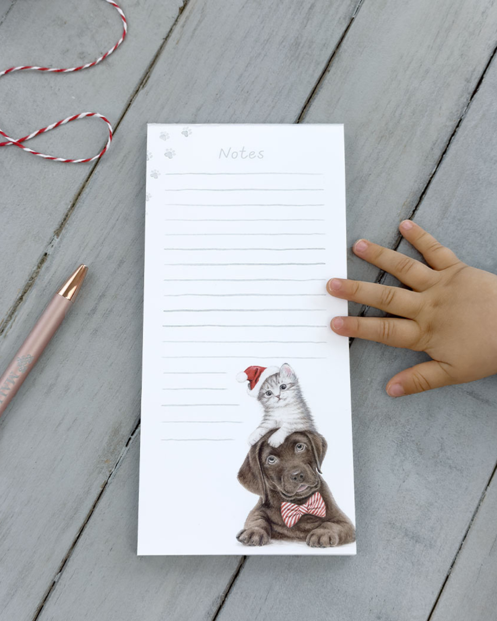 Dekorierter Maluu Notizblock Christmas Pets auf grauem Holztisch mit Kinderhand und Stift, Motiv mit Hund und Kätzchen in Weihnachtsmütze.