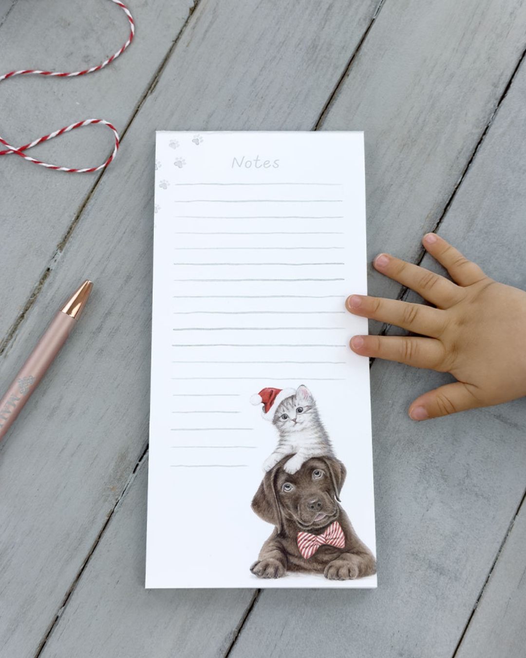 Dekorierter Maluu Notizblock Christmas Pets auf grauem Holztisch mit Kinderhand und Stift, Motiv mit Hund und Kätzchen in Weihnachtsmütze.