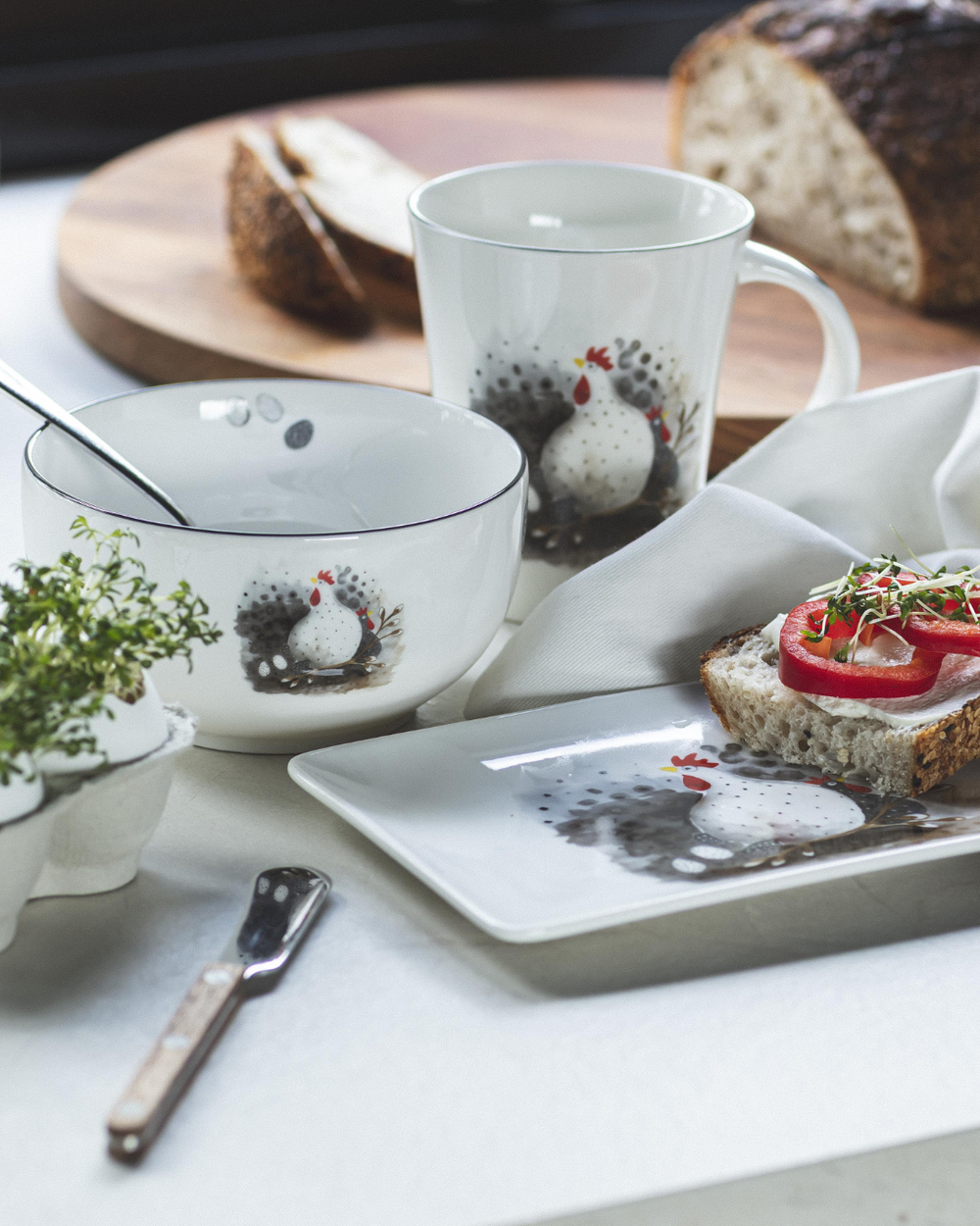 Geschirr „Höna“ von Nääsgränsgården auf einem Frühstückstisch mit Schale, Tasse, Teller und frischen Kräutern. Das Motiv zeigt Hühner und Eier, harmonisch in eine ländliche Frühstücksszene eingebettet.