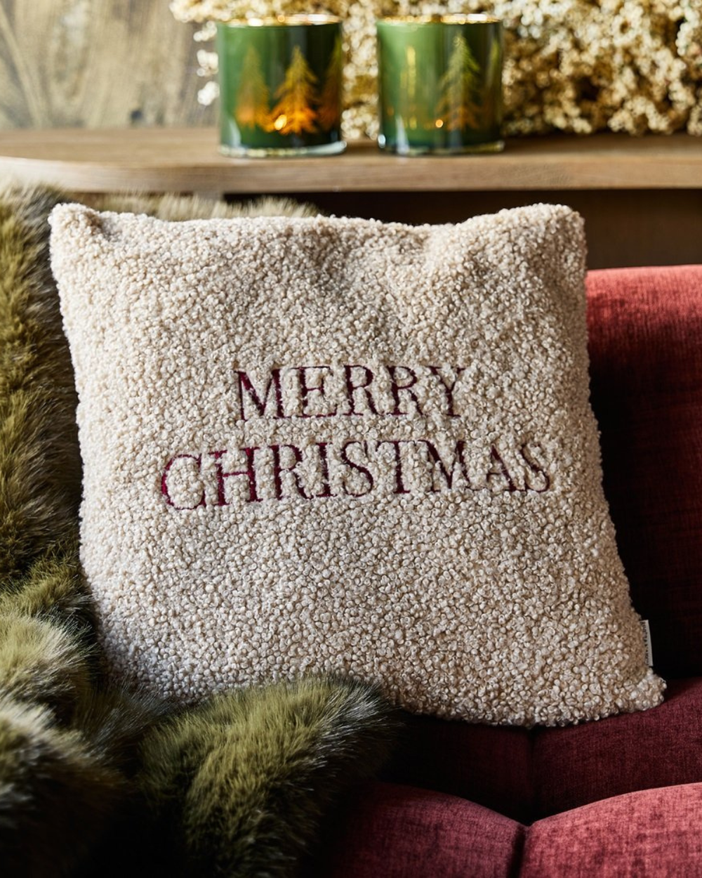 Weihnachtskissenbezug Merry Christmas aus Teddystoff dekoriert auf rotem Sofa.