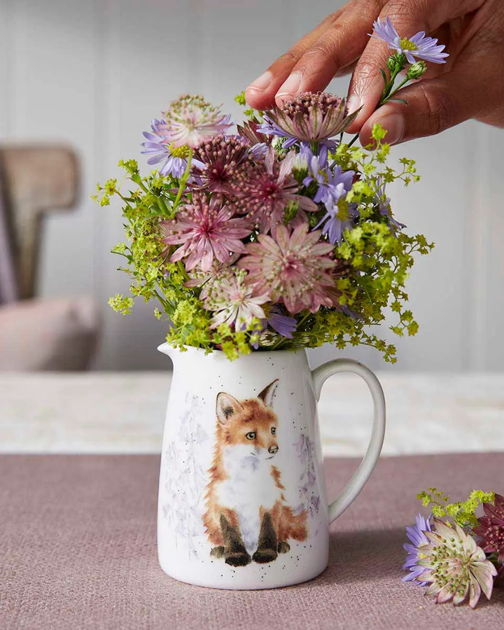 Mini Fuchs Krug von Wrendale Designs auf einem dekorierten Holztisch. Das Motiv ist ein niedlicher Fuchs mit blassen Blumen im Hintergrund. Der Krug ist mit Blumen gefüllt.