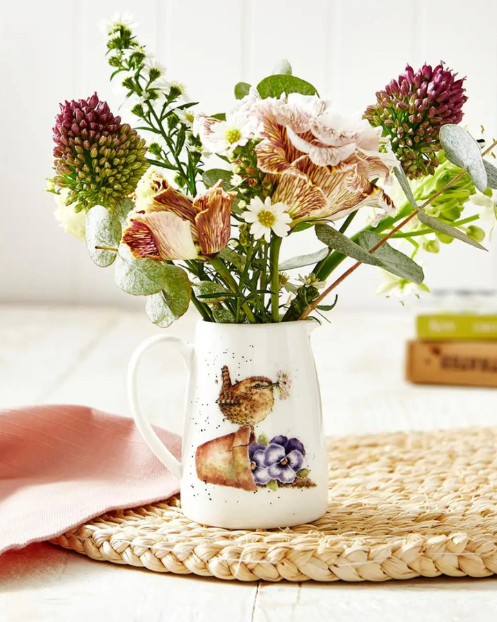 Vogel Krug von Wrendale Designs gefüllt mit Blumen Auf einem hellen Holztisch. Das Motiv des Krugs ist ein niedlicher Vogel welcher Blumen mit den Zähnen hält, während er auf einem Umgefallenen Blumentopf steht.