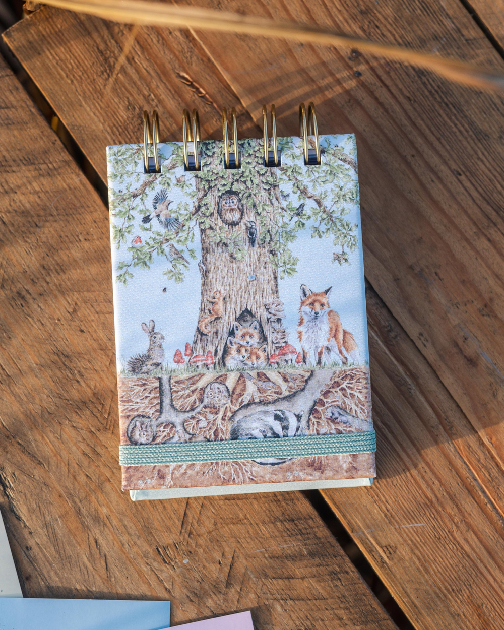 Geschlossenes Mini Notiz- und Skizzenbuch „Woodland“ mit umlaufendem Waldmotiv und grünem Gummiband auf einem Holztisch dekoriert.