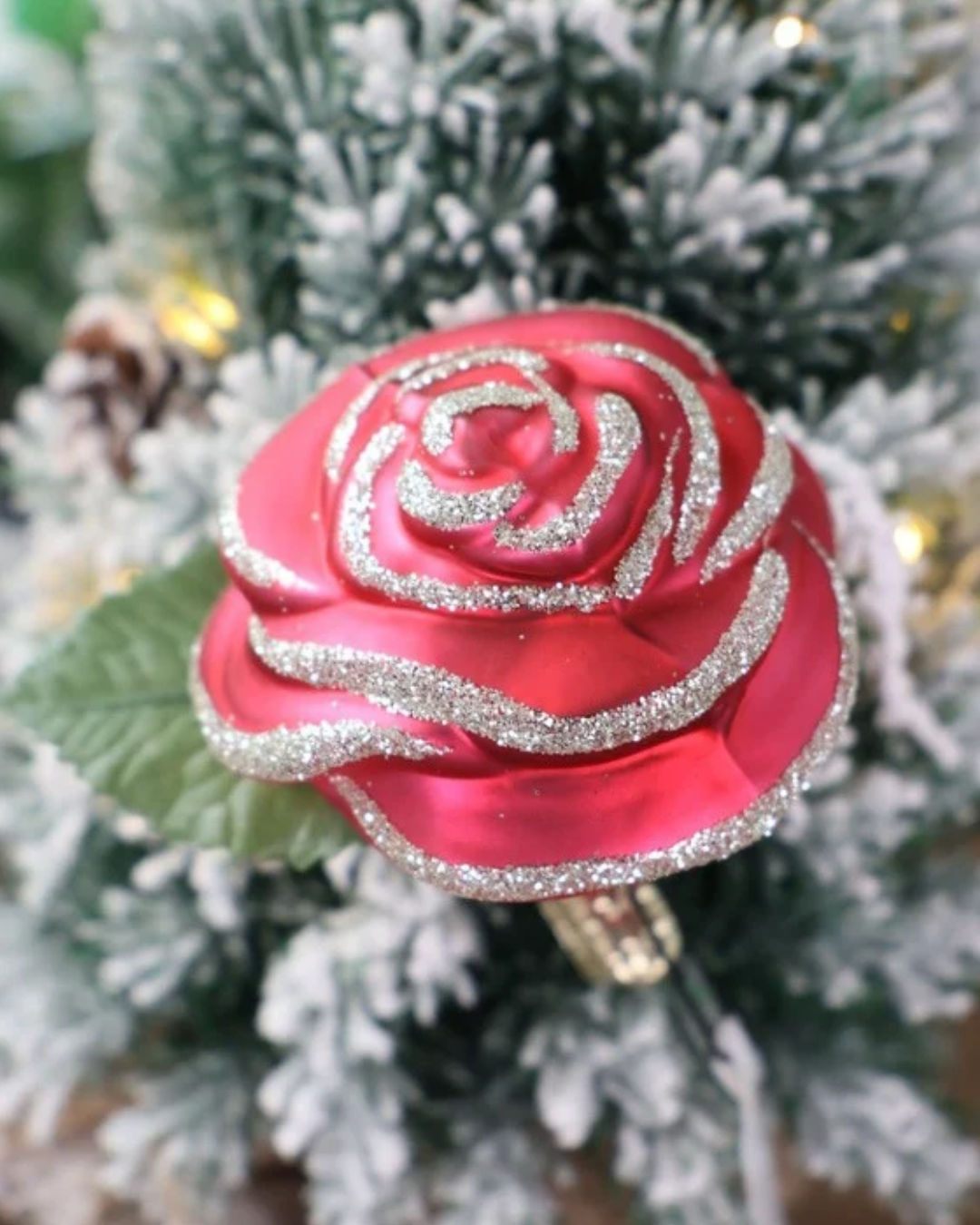 Weihnachtliches Rosen Ornament zum Klemmen in Deutschland handgefertigt von Inge Glas vor weißem Hintergrund. Die Rose hat rote Blütenblätter mit silbernen Glitzer Highlights und einem grünen Blatt unterhalb der Blüte. Hier angebracht auf einem weiß gepuderten Tannenbaum.