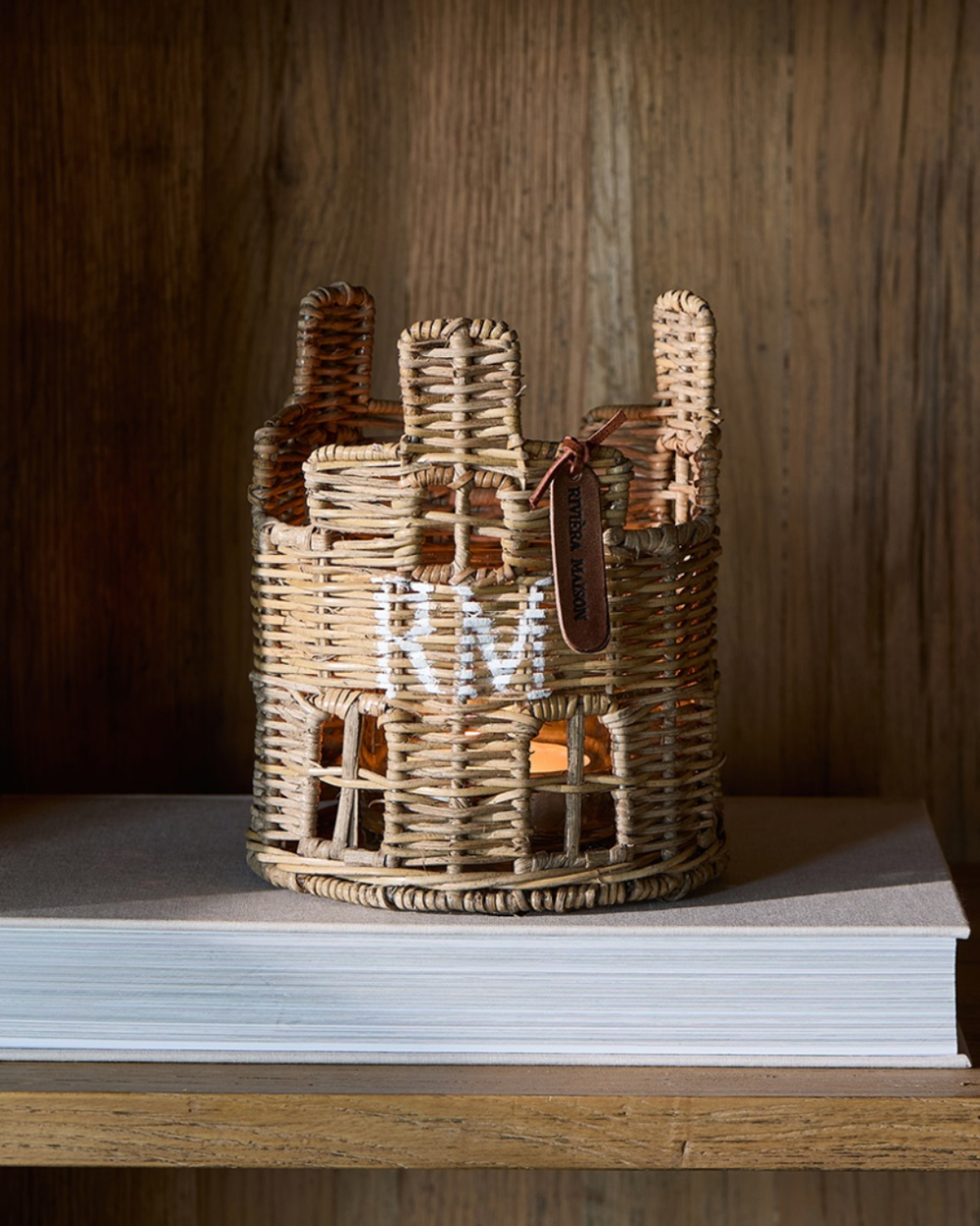 Das Rustic Rattan House Votive von Riviera Maison aus handgefertigtem Rattan steht auf einem Buch. Im Inneren des Windlichts ist eine entzündete Kerze zu sehen, welche im mitgelieferten Glas steht. Der "RM" Schriftzug in weiß ist gut auf dem Windlicht zu erkennen. Ein ledernes Schildchen Riviera Maison Aufschrift ist neben einen der drei Schornsteine geknotet.
