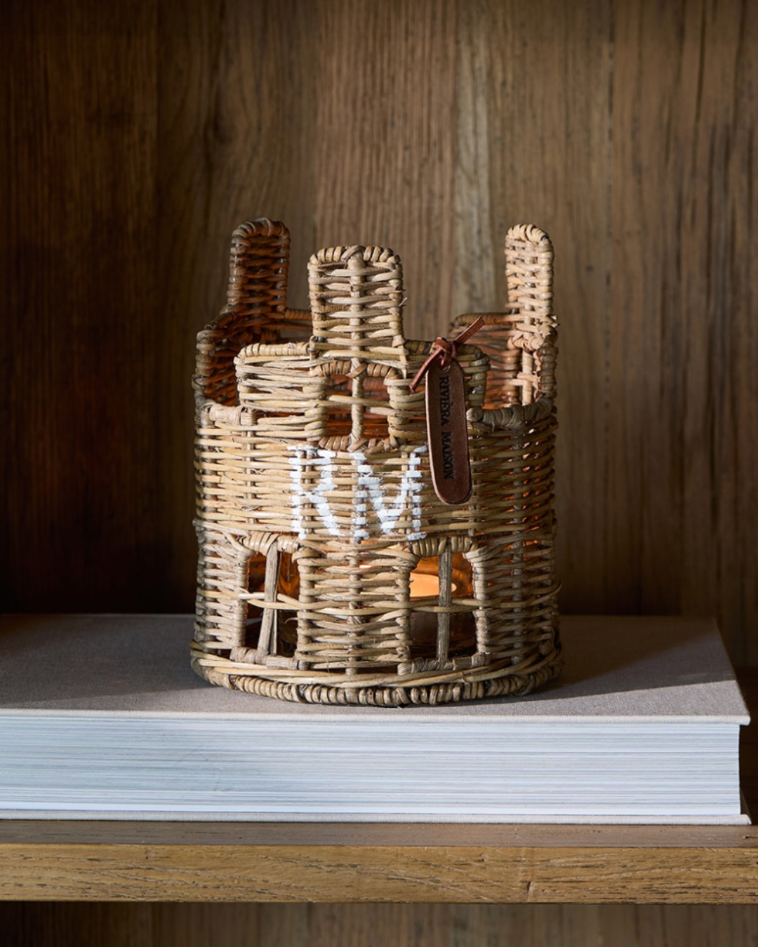 Das Rustic Rattan House Votive von Riviera Maison aus handgefertigtem Rattan steht auf einem Buch. Im Inneren des Windlichts ist eine entzündete Kerze zu sehen, welche im mitgelieferten Glas steht. Der "RM" Schriftzug in weiß ist gut auf dem Windlicht zu erkennen. Ein ledernes Schildchen Riviera Maison Aufschrift ist neben einen der drei Schornsteine geknotet.