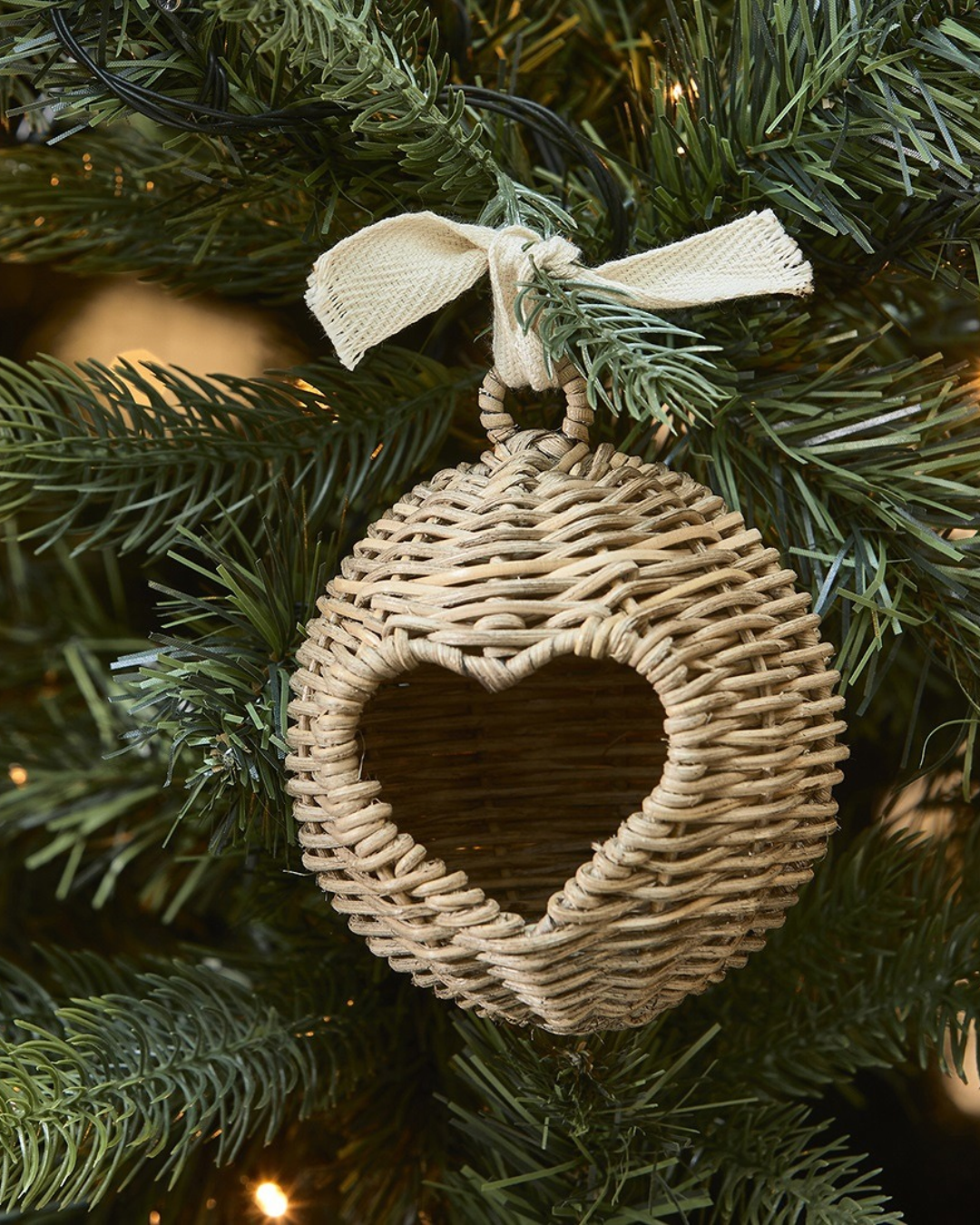 Dekoriertes "Rustic Rattan Love Bauble" Ornament von Riviera Maison. Das Ornament hängt an einem Tannenbaum mit der Herz Seite zur Kamera gedreht.