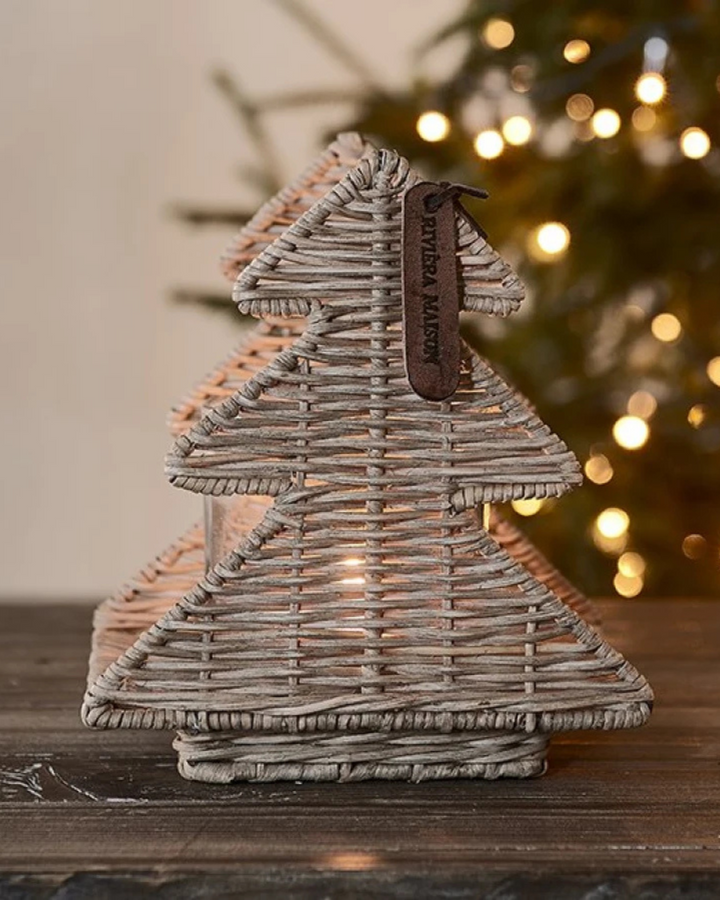 Dekoriertes Rustic Rattan Lovely Trees Windlicht von Rivièra Maison mit Kerzenschein, im Hintergrund ein Weihnachtsbaum mit Lichtern.