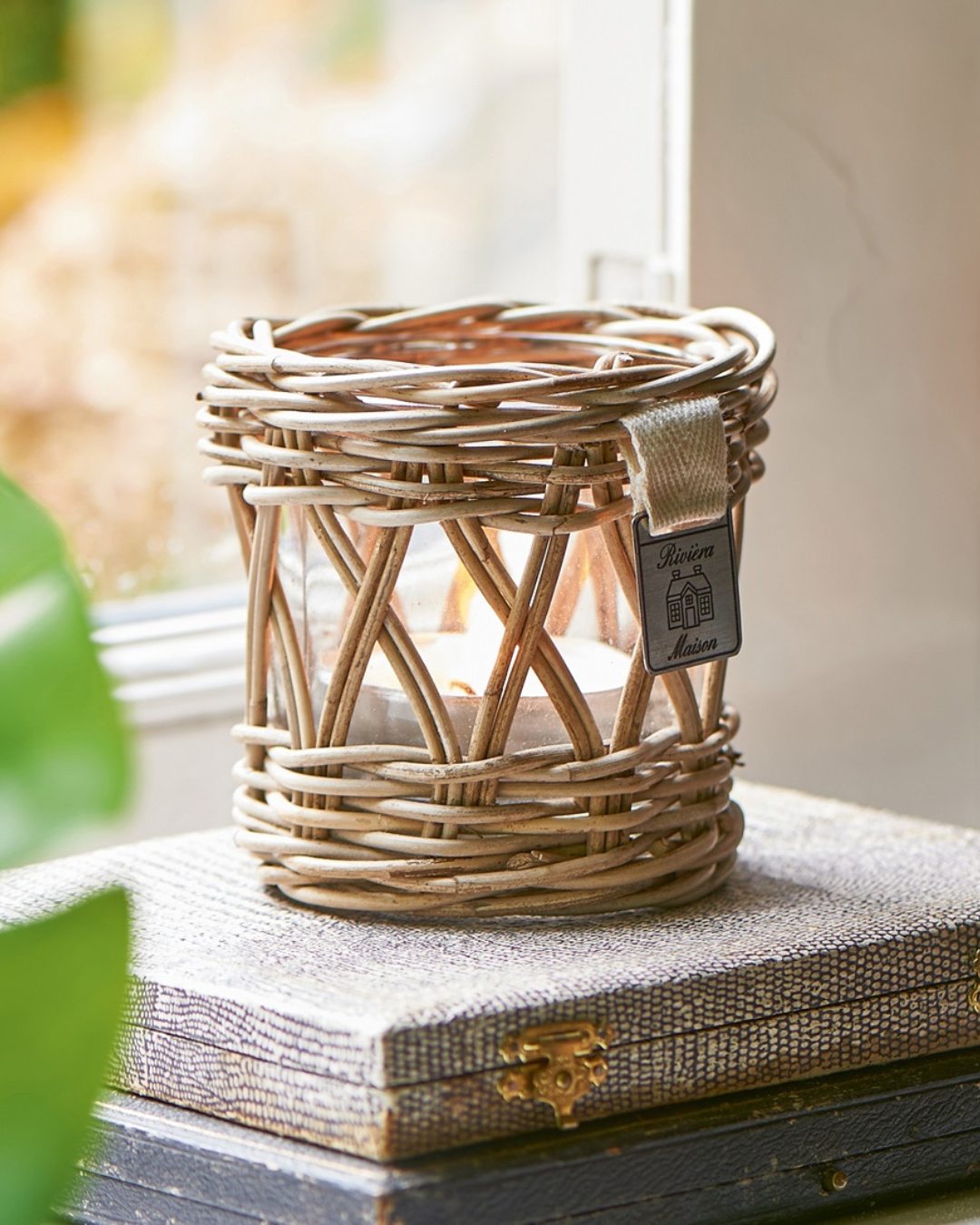 Das Rustic Rattan Votive Windlicht ist vor einem Fenster dekoriert und hat ein kleines Teelicht im Inneren des Glases. Die Dekoration von Rivièra Maison besteht aus geflochtenem Rattan und hat ein Stilvolles Muster, durch welches das Kerzenlicht scheint.