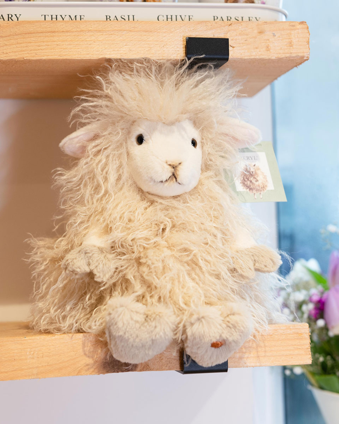Schaf Kuschelier Beryl in Größe L von Wrendale Designs sitzend auf hellen Holzregal in einem dekorierten Zimmer. Das Fell ist weiß und leicht gräulich/beige.