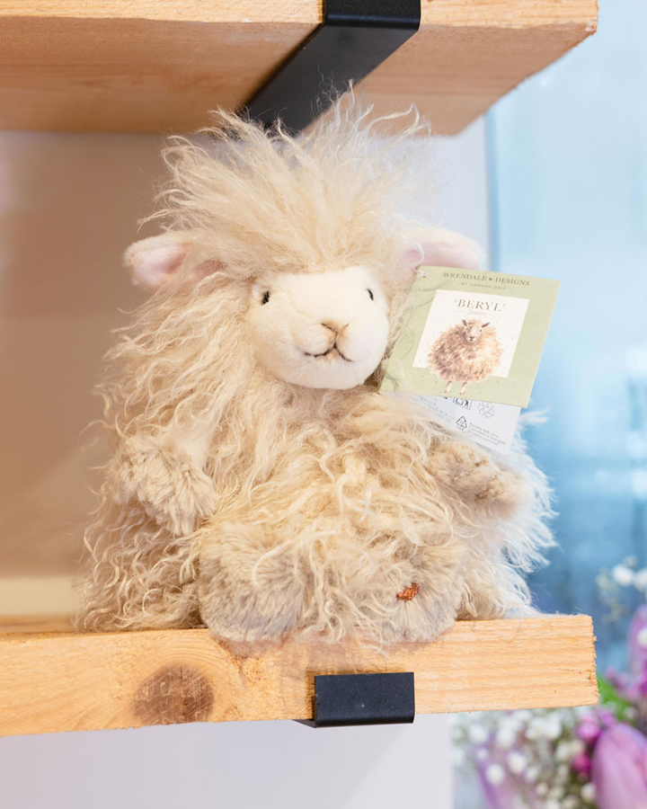 Schaf Kuschelier Beryl in Größe M von Wrendale Designs sitzend auf Holzregal in einem dekorierten Zimmer. Das Fell ist weiß und leicht gräulich/beige.