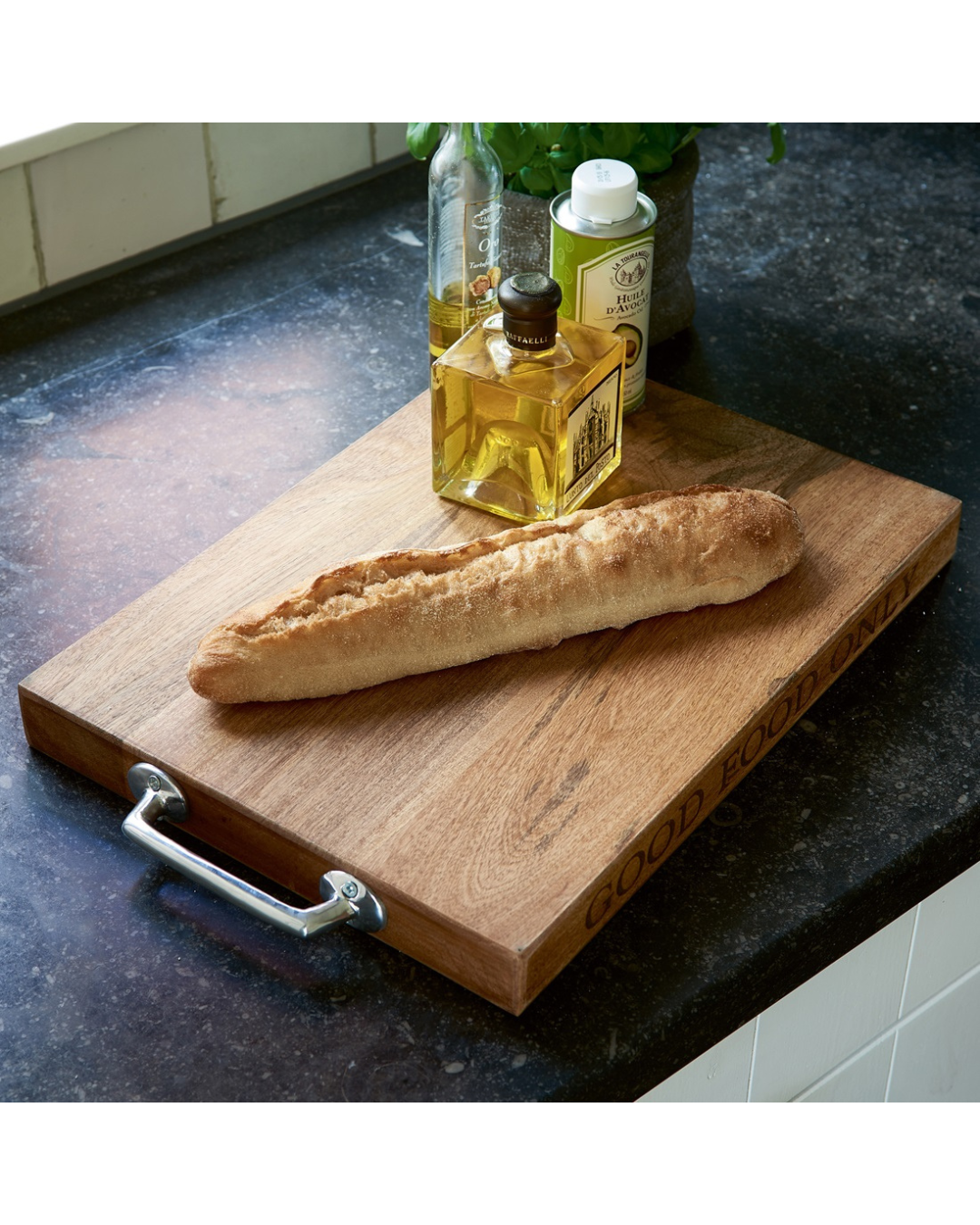 Schneidebrett „Cooking with Love“ von Rivièra Maison mit Baguette und Öl dekoriert auf einer dunklen Küchenarbeitsfläche.