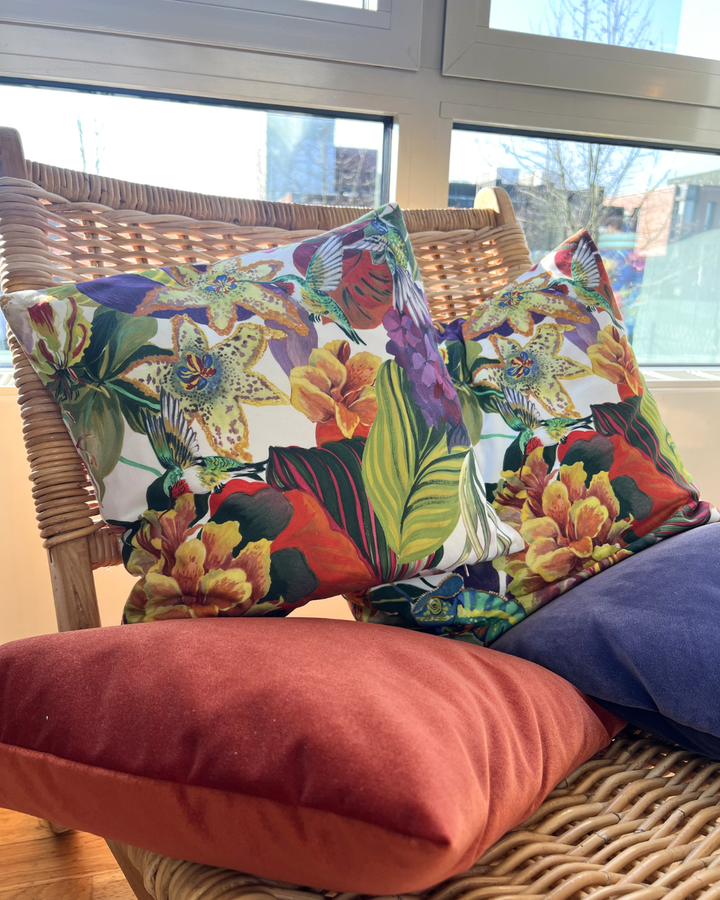 Auf dem zweiten Bild liegt der tropische Kissenbezug "Valparaiso 50x50" von Sander auf einem cremefarbenen Sofa mit weiteren Kissen in passenden Farben. Der Kissenbezug zeigt das gleiche exotische Muster aus Palmen, Blüten, Kolibris, Orchideen und einem Chamäleon.