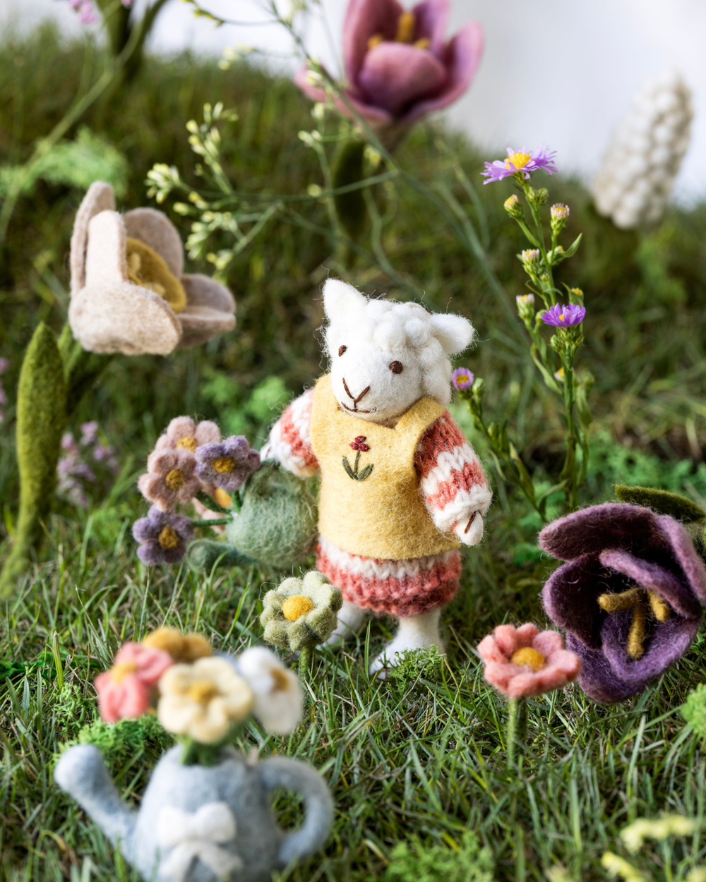 Dekoriertes weißes Filzschaf mit grüner Gießkanne von Gry & Sif in einer frühlingshaften Wiesenlandschaft mit Filzblumen.