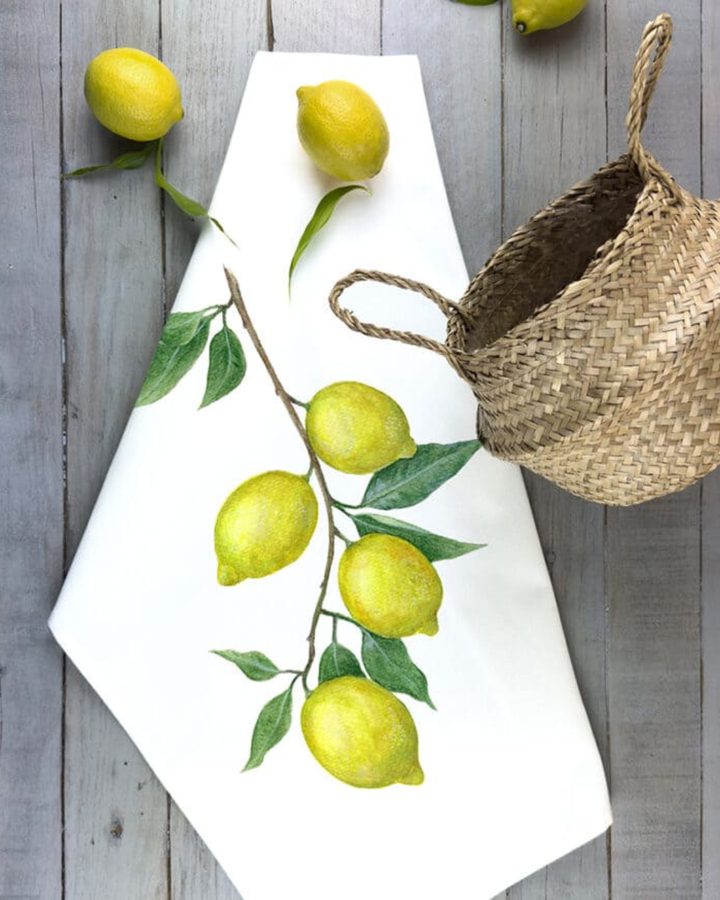 Maluu Geschirrtuch Sunny Lemons dekorativ ausgelegt mit Zitronenmotiv neben frischen Früchten und Korb auf Holzuntergrund