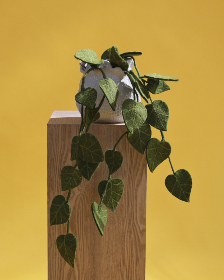 Filz Zimmerpflanze "Efeu" von Gry & Sif vor gelben Hintergrund. Die Filz Zimmerpflanze ist in einer weißen Blumenvase welche auf einem Holz Pfad steht. Die Pflanze ist gut erkennbar und ragt weit nach unten.