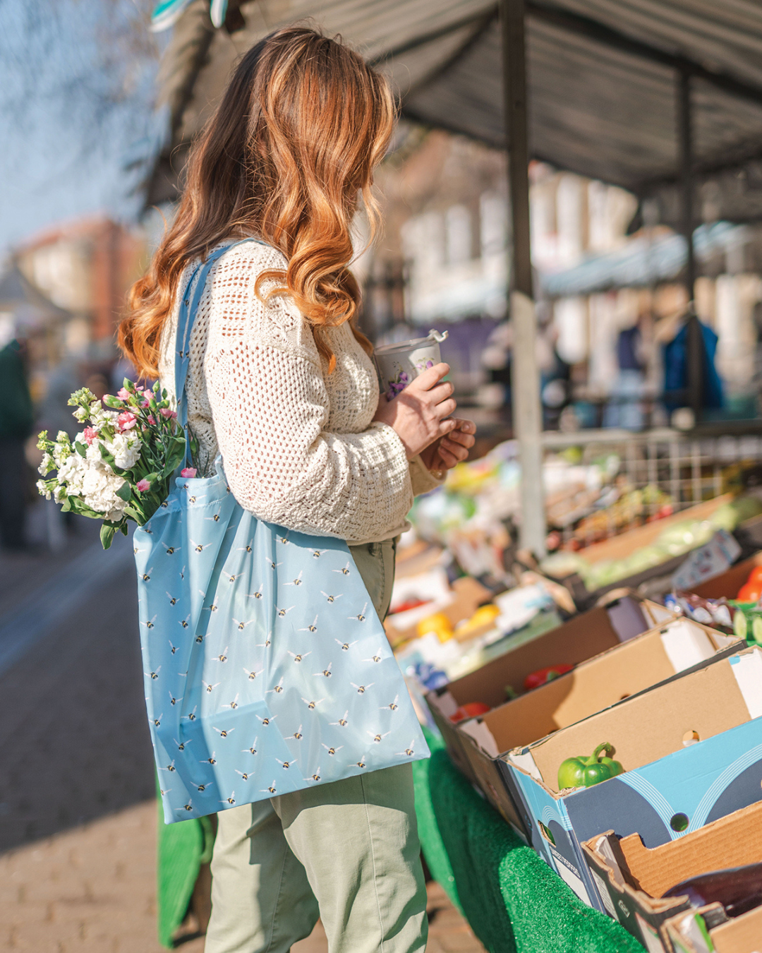 Einkaufstasche Busy Bee von Wrendale Designs im Benutzung auf dem Wochenmarkt. Eine Frau steht vor einem Gemüsestand, hält einen Becher in der Hand und trägt die Einkaufstasche über der Schuklter. Die von Hannah Dale liebevoll illustrierten Hummeln im Flug sind auf der Tasche gut erkennbar.